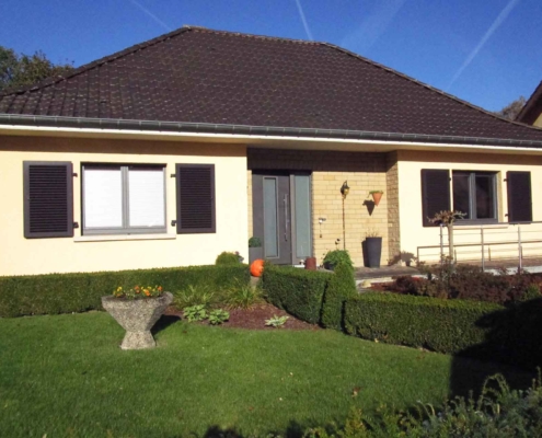 Gelbliches Einfamilienhaus mit einer modernen grauen Coplaning Keramik Haustür und braunen Coplaning Holz Jalousienfenster.