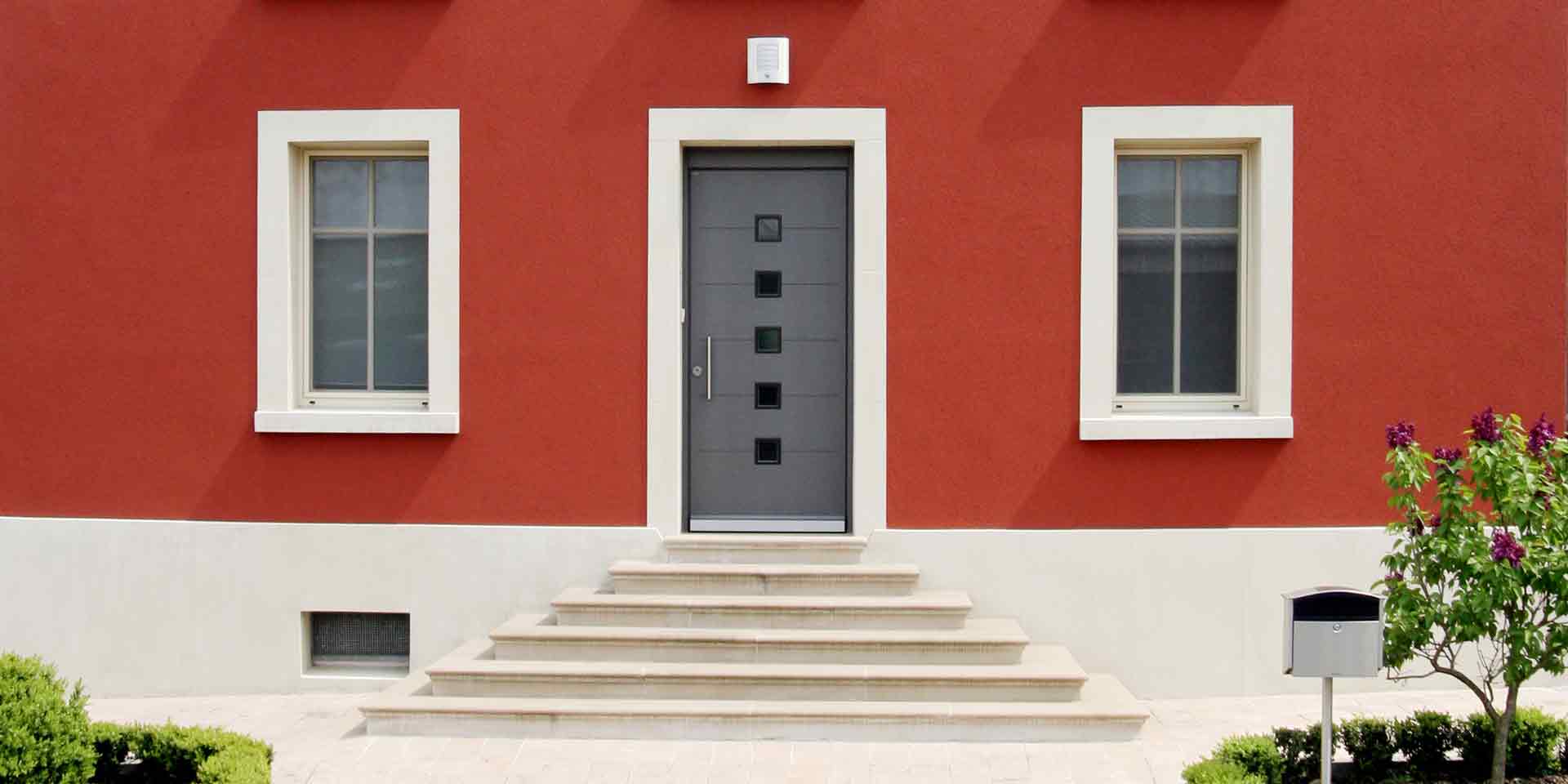 Nahaufnahme auf den Eingangbereichs des roten Einfamilienhauses mit Sicht auf die Coplaning Haustür und jeweils links und rechts davon weiße Coplaning Fenster.