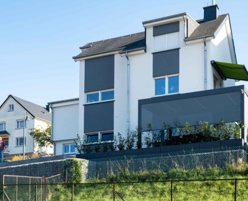 Weißes Einfamilienhaus mit Blick auf eine graue Alu Coplaning Veranda Terassenüberdachung mit Lamellendach und kippbarem Lamellendach mit LED-Licht.