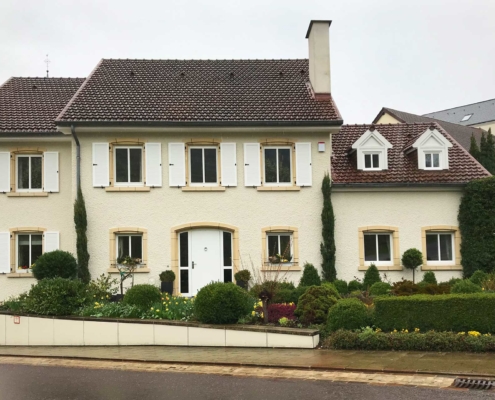 Gelbliches Einfamilienhaus mit moderner weißer Coplaning Haustür und weiße Coplaning Holzfenster mit weißen Klappläden.